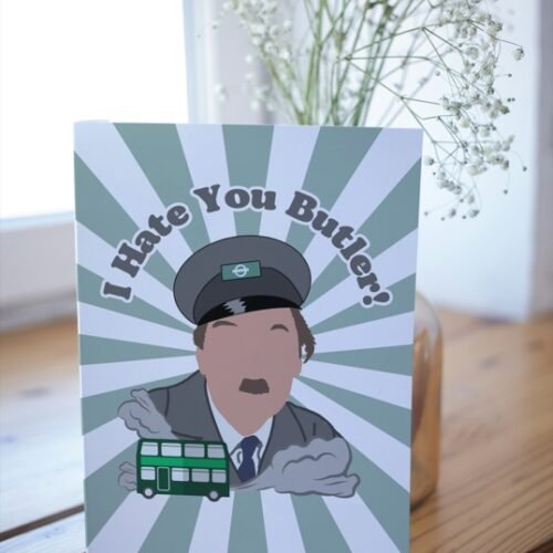 Screenshot of a customizable greeting card featuring the playful quote 'I hate you butler!' in bold, retro-style font, referencing the classic British sitcom On the Buses. The text is set against a plain or lightly textured background, evoking a humorous and nostalgic tone for fans of the show. This features the card upright on a table with a cartoon of character from the show Blake.