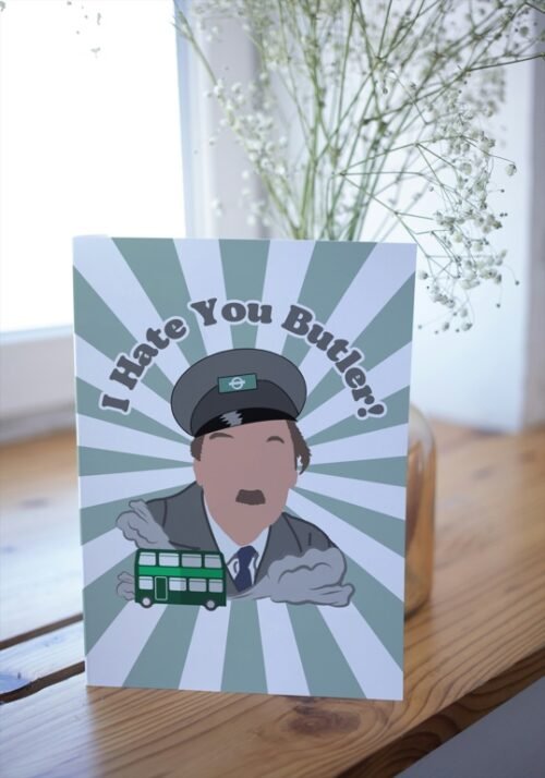 Screenshot of a customizable greeting card featuring the playful quote 'I hate you butler!' in bold, retro-style font, referencing the classic British sitcom On the Buses. The text is set against a plain or lightly textured background, evoking a humorous and nostalgic tone for fans of the show. This features the card upright on a table with a cartoon of character from the show Blake.