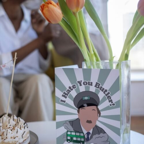Screenshot of a customizable greeting card featuring the playful quote 'I hate you butler!' in bold, retro-style font, referencing the classic British sitcom On the Buses. The text is set against a plain or lightly textured background, evoking a humorous and nostalgic tone for fans of the show. This features the card upright on a table with a cartoon of character from the show Blake.