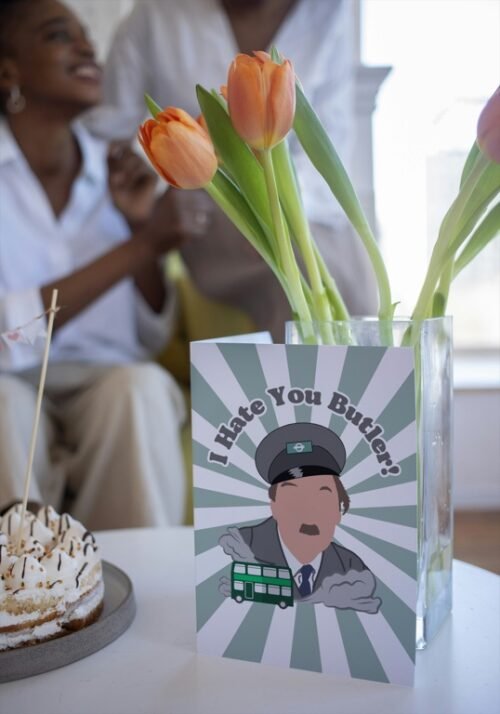 Screenshot of a customizable greeting card featuring the playful quote 'I hate you butler!' in bold, retro-style font, referencing the classic British sitcom On the Buses. The text is set against a plain or lightly textured background, evoking a humorous and nostalgic tone for fans of the show. This features the card upright on a table with a cartoon of character from the show Blake.